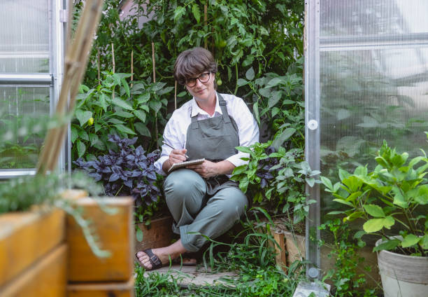 A joyful mid-adult Caucasian woman enjoys tending to her plants in a greenhouse, dressed in casual work attire with apron, holding a notebook.