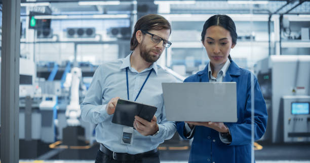 istock Caucasian Engineer and Asian Female Project Manager Standing in Office, Talking and Using Computers and Monitoring Production at Modern Automated Electronics Manufacture with AI Robotic Arms 2167547066