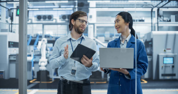 istock Caucasian Engineer and Asian Female Project Manager Standing in Office, Talking and Using Computers and Monitoring Production at a Modern Automated Electronics Manufacture with AI Robotic Arms 2167547081
