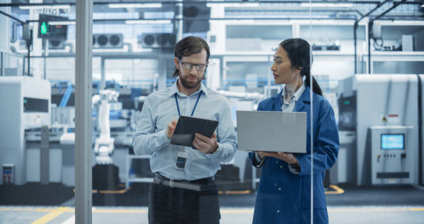 istock Caucasian Engineer and Asian Female Project Manager Standing in Office, Talking, Using Computers and Monitoring Production at a Modern Automated Electronics Manufacture with AI Robotic Arms 2167547094