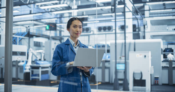 istock Electronics Manufacturing Technician in a Blue Coat Working on Laptop Computer at an Assembly Plant. Asian Female Specialist Developing, Maintaining, Setting Up Software for Automated Production 2167547130