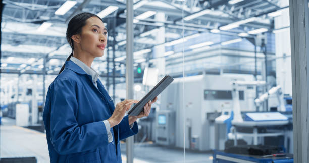 Focused Asian Engineer Monitoring and Analyzing Conditions at a Modern Electronics Factory on Tablet Device. Facility with Automated Robot Hands Working with the Help of Artificial Intelligence