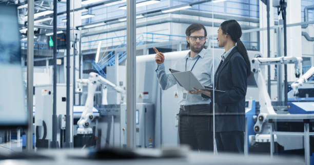 istock Portrait of a Young Engineer and Happy Asian Project Manager Using Laptop and Tablet Computers, Talking in a Factory Facility with Automated Robots Producing Modern Electronic Components 2167548603