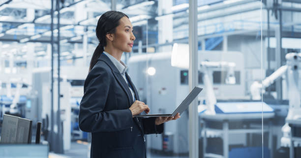 istock Production Line Technician Working on a Laptop Computer at Factory Office. Asian Female Software Specialist Updating Code for Machine Production and Assembly with Automated Robotic Arms 2167548789