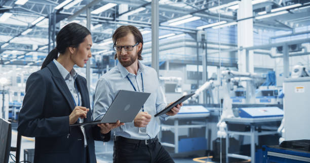 istock Male Engineer and Female Project Manager Standing in Office, Talking, Using Computers and Monitoring Production at Modern Automated Electronics Manufacture with AI Robotic Arms 2167548904