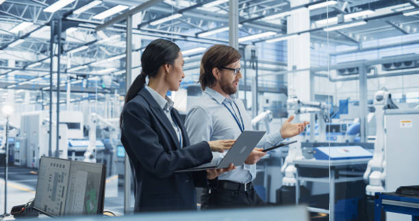 istock Male Engineer and Female Project Manager Standing in Office, Talking, Using Computers and Monitoring Production at a Modern Automated Electronics Manufacture with AI Robotic Arms 2167548936