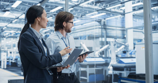 istock Portrait of Cheerful Engineer and Happy Asian Scientist Using Laptop and Tablet Computers, Talking in a Factory Facility with Robots Producing Modern Electronic Components for Different Industries 2167548956