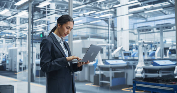 istock Beautiful Asian Female Engineer Standing Next to a Window in Office, Using Laptop Computer at Electronics Factory. Machines are Undergoing Maintenance, Specialist Using an Online Software 2167549069