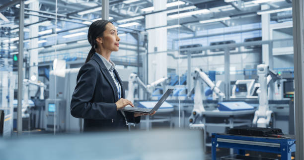 istock Beautiful Asian Female Engineer Standing Next to a Window in Office, Using Laptop Computer at an Electronics Factory. Machines are Undergoing Maintenance, Specialist Using an Online Software 2167549086