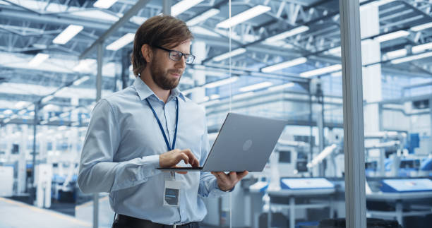 istock Portrait of a Handsome Engineer with Glasses Using Laptop Computer and Looking Around a Factory Facility with Equipment Producing Modern Electronic Components for Different Industries 2167549176