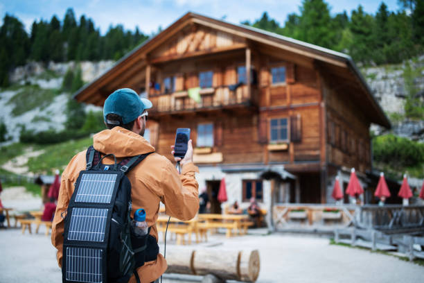 Hiker stands confidently on roof of wooden cabin, surrounded by breathtaking mountain landscape. Snow-capped peaks in distance, framed by dense evergreen forests.