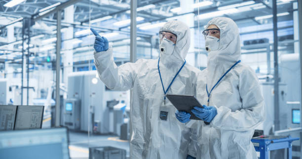 istock Bioengineers in Protective Coveralls, Masks and Eyewear Using Tablet Computer at a Chemical Manufacturing Complex. Laboratory Specialists Monitoring Conditions at a Modern Factory with Robotic Arms 2167551975