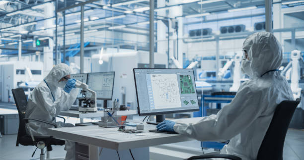 istock Establishing Shot of Two Laboratory Workers in Protective Coveralls Developing a New Advanced Central Processing Unit. PCB Design Engineer Working on a Computer, While Colleague is Using a Microscope 2167552057