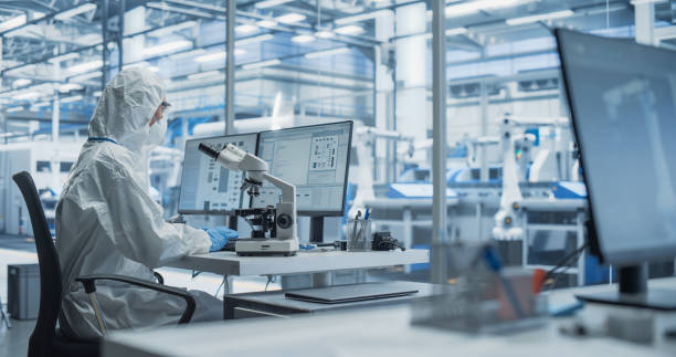 modern factory: industrial engineer working on desktop computer in clean sterile coveralls. scientist using a microscope, developing advanced solutions for high tech medical vaccine and gene research - assemblagelijn fotos stockfoto's en -beelden