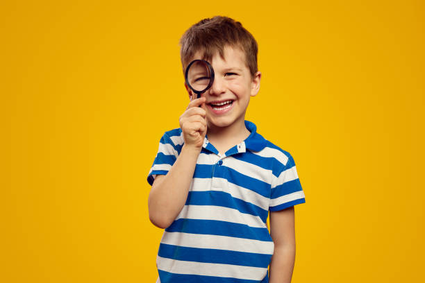 istock Cute curious kid in casual wear looking at camera through magnifier while standing against yellow 2167553462