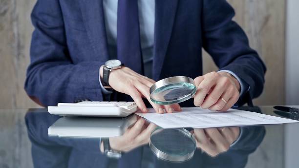 istock Auditor man analyzing financial documents with magnifying glass and calculator at his glass office desk in formal blue jacket. Business people concept 2167567146