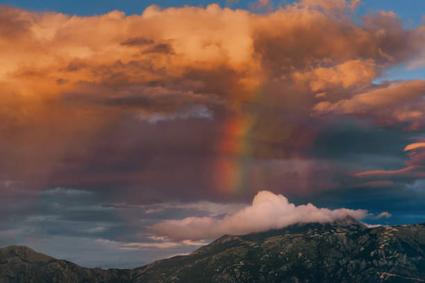 Rainbow among bright dramatic clouds softly glowing orange yellow colors from setting sun over mountains. Alpine landscape with vibrant rainbow over mountains after rain. Beauty of nature concept.