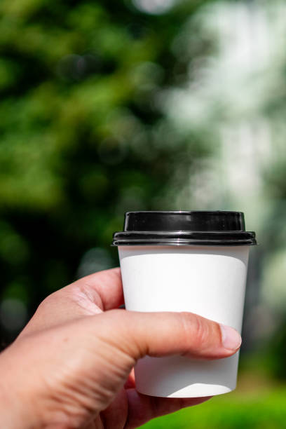 istock A hand holding a white takeaway coffee cup with a black lid, set against a blurred green background. Ideal for themes of coffee, takeaway, nature, and lifestyle. 2167568956
