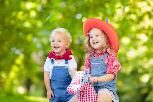 Little boy and girl dressed up as cowboy and cowgirl playing with toy rocking horse in park. Kids play outdoors. Children in Halloween costumes at trick or treat. Toys for preschooler or toddler child