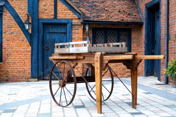 A vintage wooden cart with large iron wheels stands on a paved area in front of a quaint brick cottage. The cart, featuring a simple design with a weathered crate on top, evokes a sense of rural charm and historical craftsmanship. The cottage behind it, with its blue wooden door and leaded glass windows, adds to the rustic ambiance, highlighting the traditional architecture and timeless appeal of the scene.