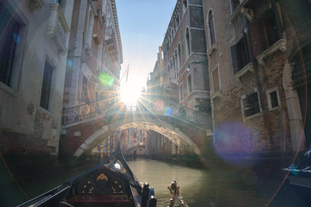 A B&W shot of a Venetian gondola silhouette and its operator crossing Venice's waters below a bridge.