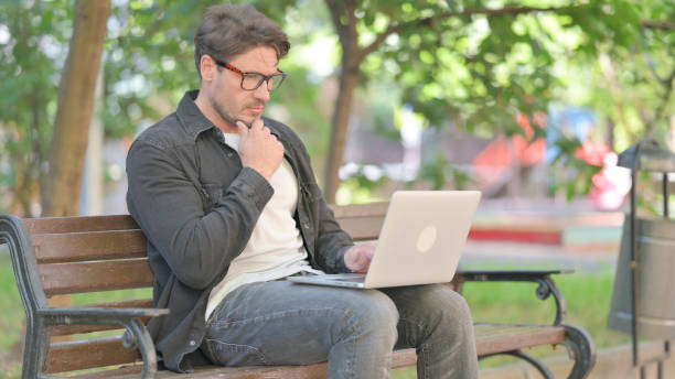 Pensive Middle Aged Man Reading on Laptop while Sitting Outdoor on Bench