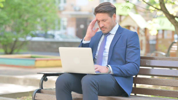 istock Businessman with Migraine Using Laptop while Sitting Outdoor on Bench 2167616409