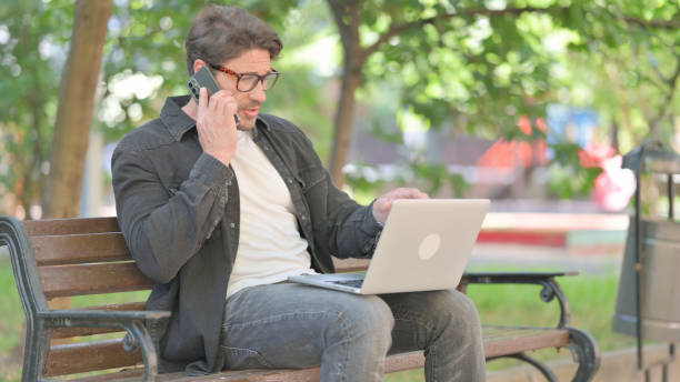 istock Middle Aged Man Talking on phone and Using Laptop Outdoor 2167618374