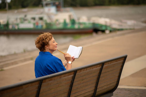 A woman sits on a bench, reading and writing with a pencil, enjoying the peaceful riverside atmosphere on a sunny afternoon.