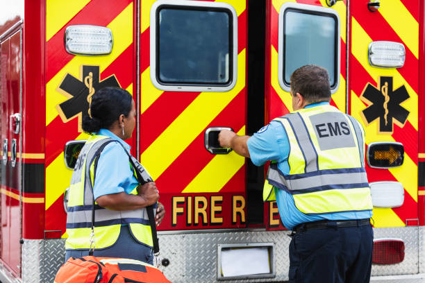 Two multiracial paramedics standing at the rear of a rescue ambulance. The Hispanic woman, in her 40s, is carrying a first responder bag, watching as her partner, a mature man in his 50s, opens the doors of the vehicle.