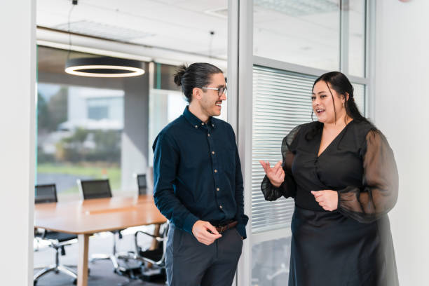 Two colleagues standing and having a discussion in a modern office space with a conference room in the background.