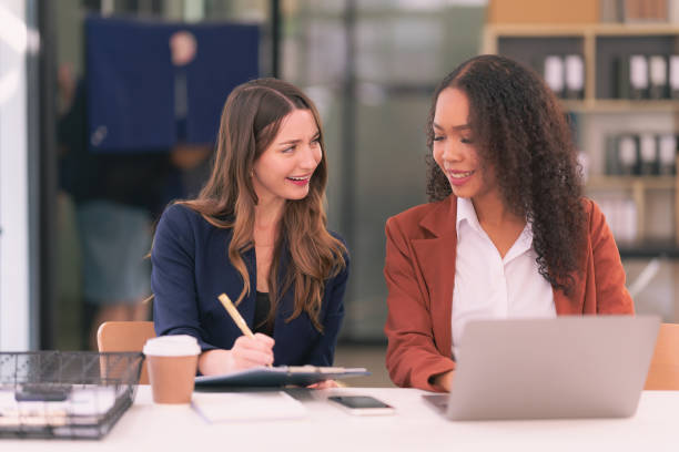 istock Two young businesswomen work together on laptops while talking about panning, reading documents and writing notes while discussing a new project. 2167703405