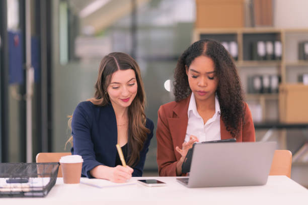 istock Two young businesswomen work together on laptops while talking about panning, reading documents and writing notes while discussing a new project. 2167703420