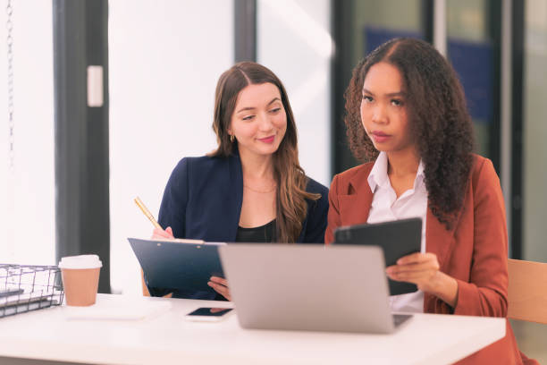 istock Two young businesswomen work together on laptops while talking about panning, reading documents and writing notes while discussing a new project. 2167703421
