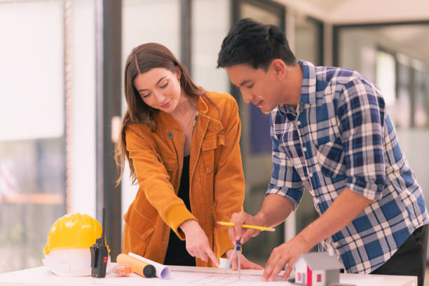 istock Two male and female architects designing a construction project on blueprints and a house model standing in the office are talking about placing a real estate business girlfriend. 2167703466
