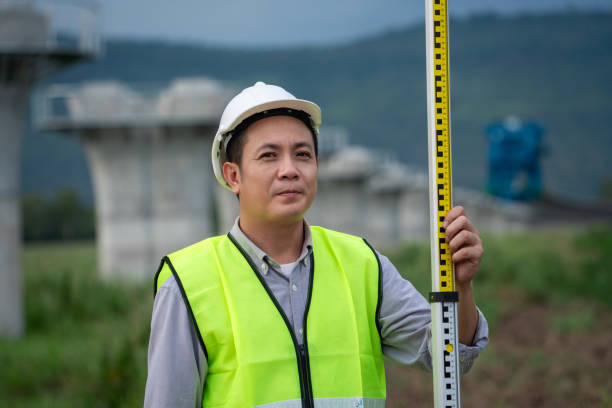 A man in a yellow vest is holding a yellow tape measure. He is wearing a hard hat and is standing in a field