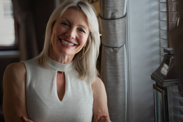 A mature woman is standing next to a window in a hotel lounge, she is laughing and is wearing an off white sleevless top. Her short hair is blonde.  Horizontal image.