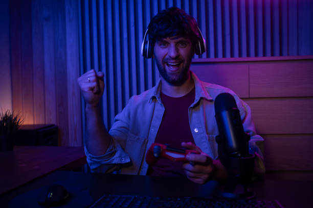 istock A gamer is fully immersed in an engaging session while holding a controller. The room is illuminated with colorful lighting, creating a vibrant atmosphere. The microphone positioned nearby captures every moment of excitement and commentary. 2167736686