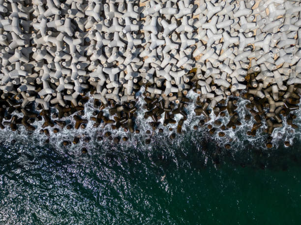 Aerial view of concrete tetrapod wave breakers along a rugged shoreline, with waves crashing against them, creating a dynamic and textured seascape. Ideal for coastal, industrial, and nature themes