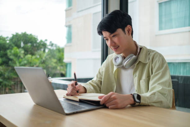 istock Young man is sitting at a wooden table, focused on his work with a laptop and a notebook. 2167782000