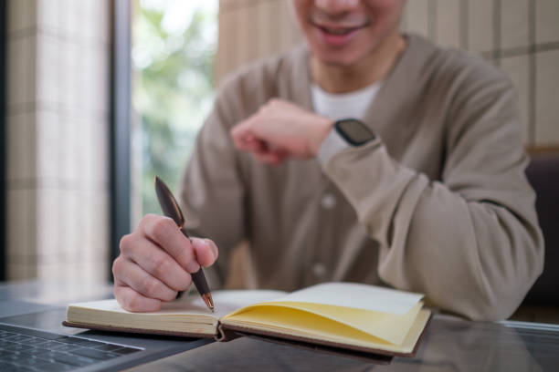 This close-up image captures a man writing in a notebook, focusing on his hand holding a pen and the notebook on a table.