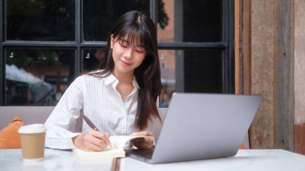 istock A young professional woman sits at a table, writing in a notebook while working on her laptop. 2167782413