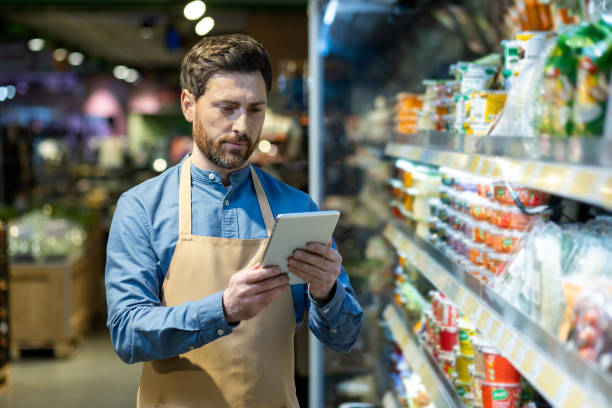istock Grocery store worker wearing apron using tablet for inventory management in food aisle 2167787657