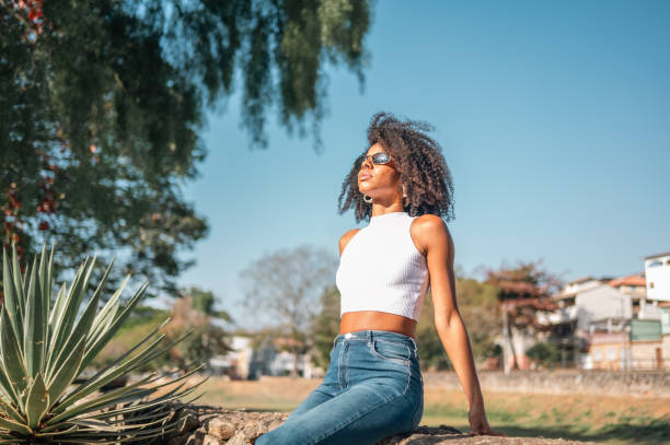 Dressing in a strapless colorful dress, wearing a golden neck ring, a pretty black woman with ponytail hair is standing at small woods inside a big city, looking around, waiting for you.\