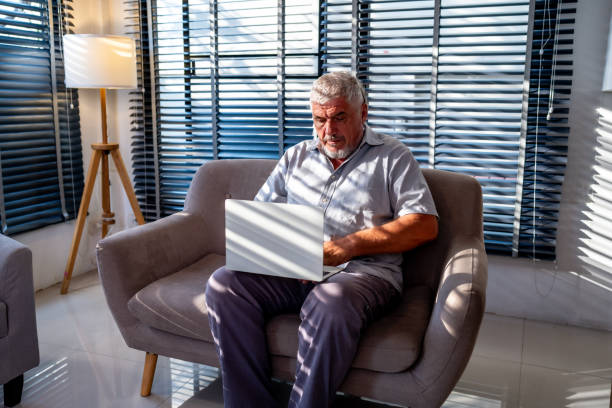 istock Elderly man sitting on a sofa using a laptop in a bright, modern living room with sunlight streaming through window blinds. 2167801275