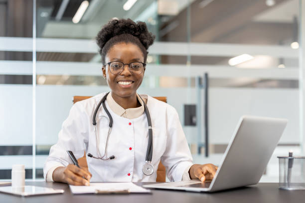 istock Smiling doctor working at desk with laptop and writing notes in modern office 2167808044
