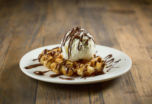 plate of belgian waffle with vanilla ice cream and melted chocolate sauce on wooden table