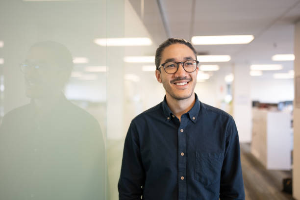 Portrait Photo of Smiling Asian Male Portrait photograph of a relaxed Asian office worker standing next to a reflective glass panel in a corporate office man standing profile stock pictures, royalty-free photos & images