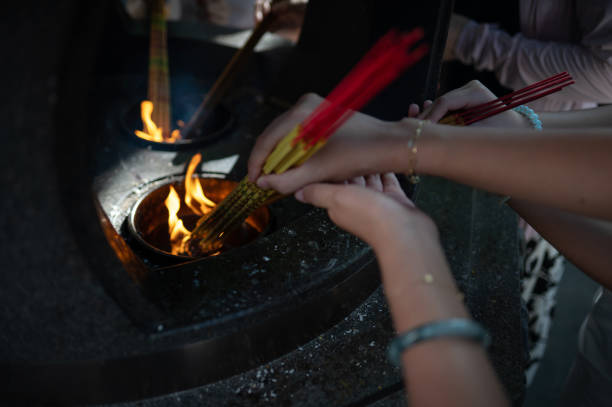 Participants light incense sticks from flames as part of a traditional ritual. The scene features hands holding incense sticks near an open flame, with a stone structure providing the ambiance for the religious or cultural ceremony.