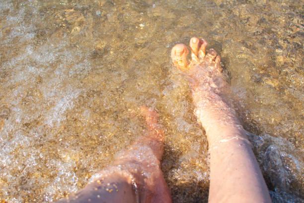 a woman is standing in the water, only her legs are visible, transparent clear water, pebbles and the reflection of the sun are visible
